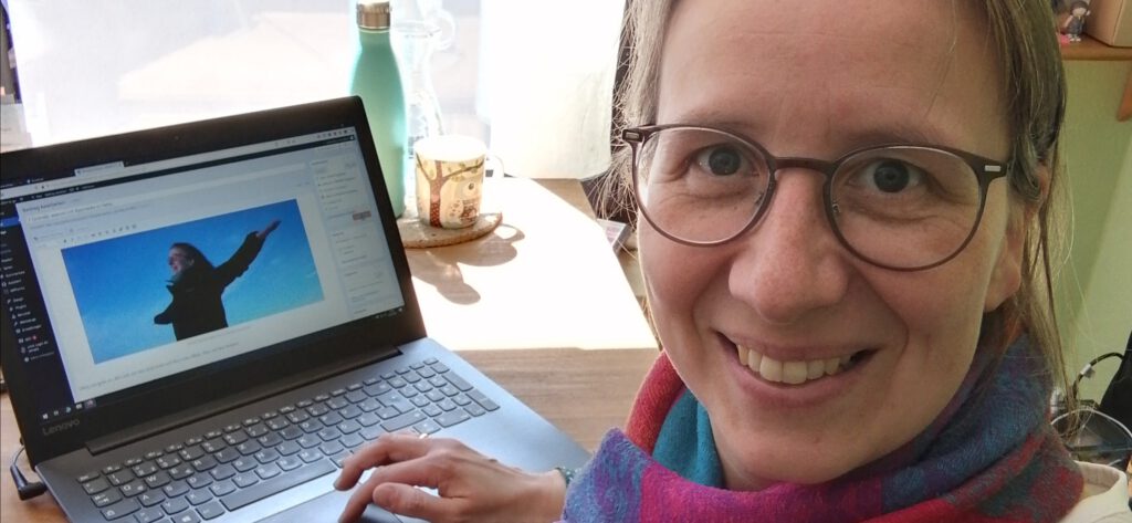 Bernadette sitzt an ihrem Schreibtisch und strahlt glücklich in die Kamera, weil sie gerade an einem Blogartikel schreibt.
