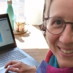 Bernadette sitzt an ihrem Arbeitsplatz, einem Hochtisch mit Laptop und warmem Wasser
