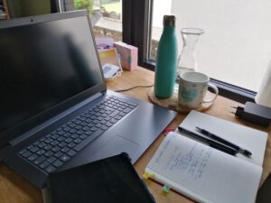 Schreibtisch mit Laptop, Kalender, bulletJournal und Bernadettes Thermoskanne, einer Wasserkaraffe und einer Tasse.