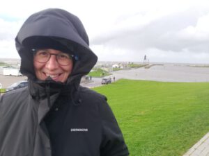 Bernadette steht mit Regenmantel, Mütze und Kapuze am überfluteten Nordsee-Strand und strahlt in die Kamera.