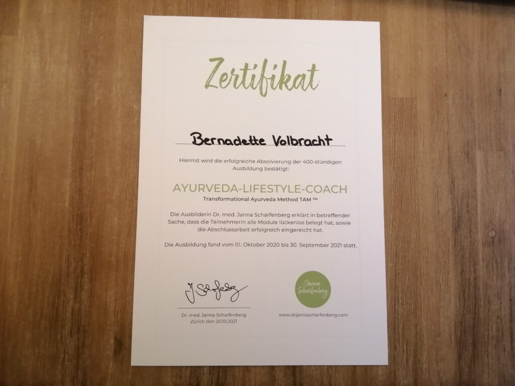 Bernadettes Zertifikat zum Ayurveda-Lifestyle-Coach liegt auf einem Holztisch.