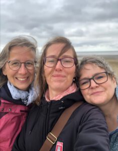 Bernadette, Kerstin und Anja stehen aneinandergekuschelt am Strand. Die Haare vom Wind zersaust, alle strahlen in die Kamera.
