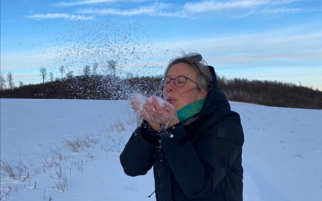 BErnadette steht auf einer verschneiten Wiese und pustet Schnee aus ihrer Hand. Dieser wirbelt durch die Luft.