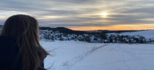 Bernadette steht mit dem Rücken zur Kamera vor einem Sonnenuntergang. Die Wiesen vor ihr sind schneebedeckt.