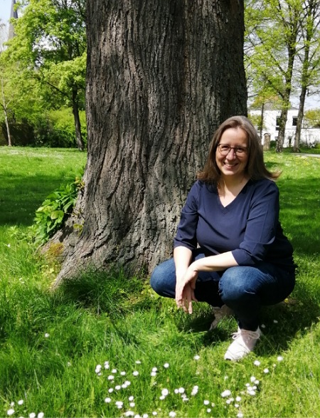 Bernadette, Ayurveda-Lifestyle-Coach, kniet vor einem alten Baum auf einer grünen Wiese.